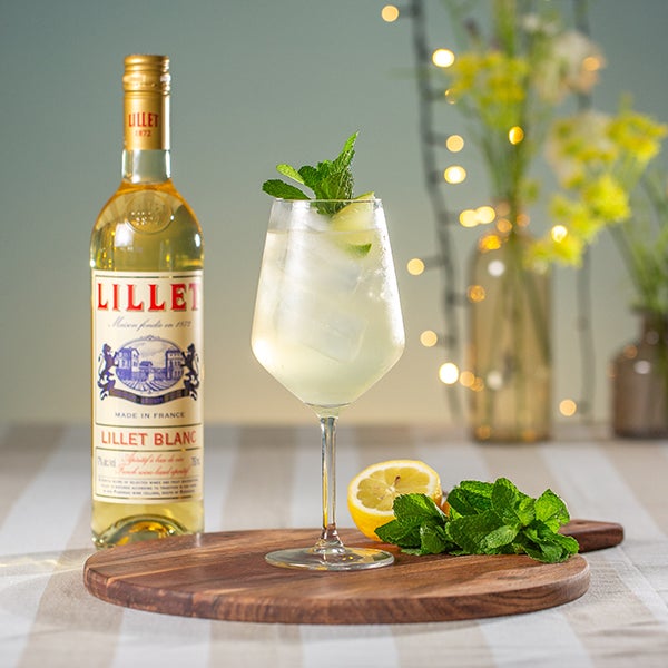 Lillet Hugo cocktail på et bord med en flaske Lillet Blanc og en buket blomster.