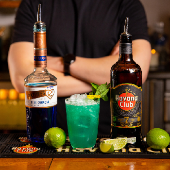 En blå Curacau flaske, en Havana club rom 7 års flaske, og en blå drink på et bord.