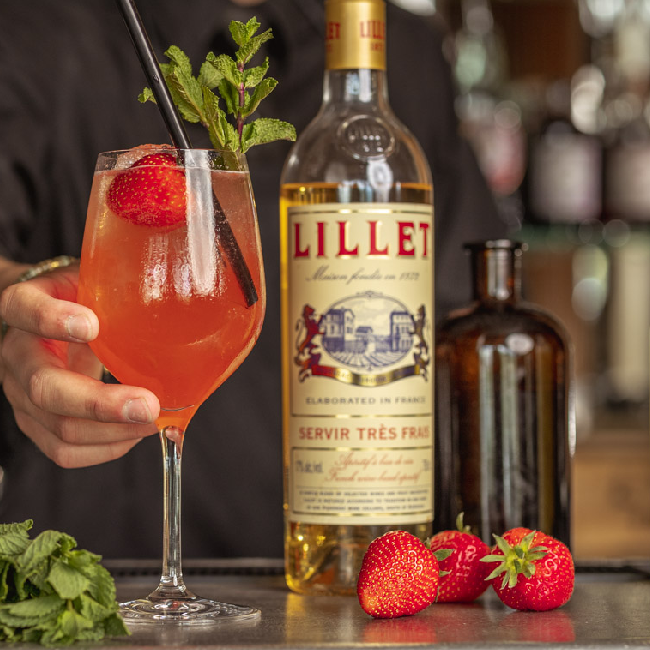 Summertime Madness cocktail på et bord med en flaske Lillet Blanc og jordbær