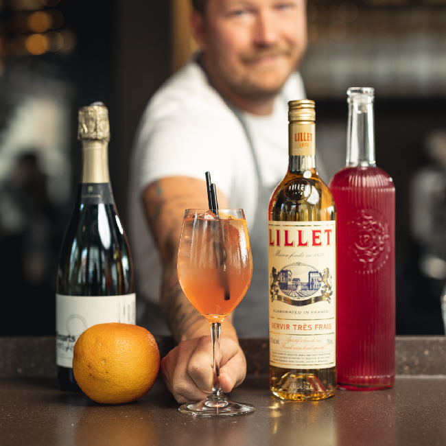 Nærvær Rabarber Spritz drink serveret i et vinglas med en flaske Lillet blanc og champagne.