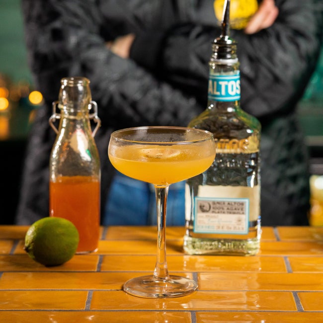Mango Margarita cocktail på et bord med en flaske Altos Plata og en lime.