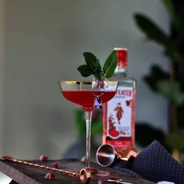 Fragaria Valentina cocktail på et skærebræt med roser, barske og en flaske Beefeater Pink Strawberry Gin