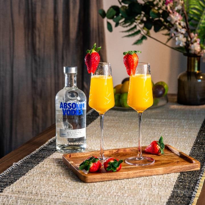 To Absolut Mimosa drinks på et skærebræt med jordbær og en flaske Absolut Vodka.