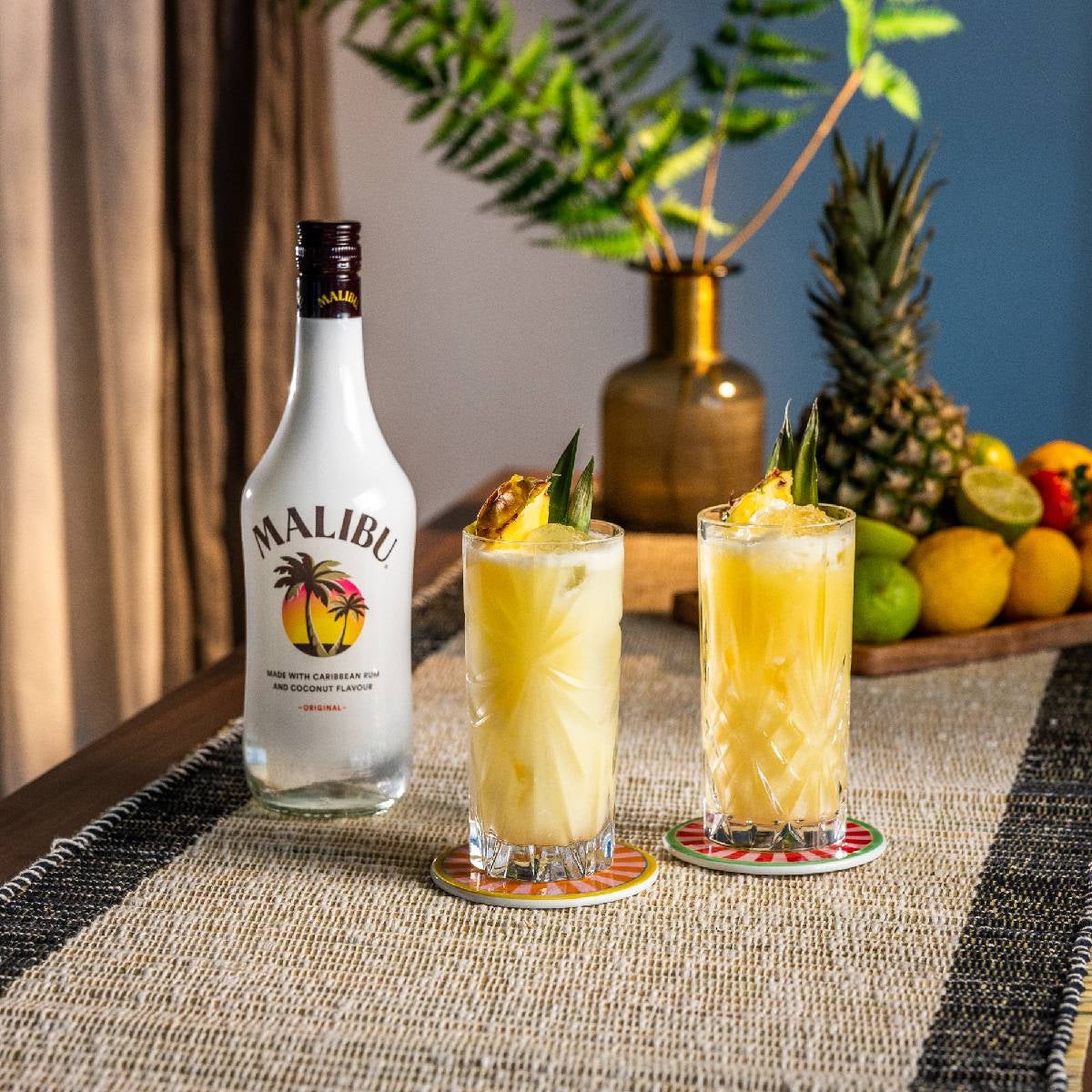 To Piña Colada cocktails på et bord med en flaske Malibu rom og frugter.