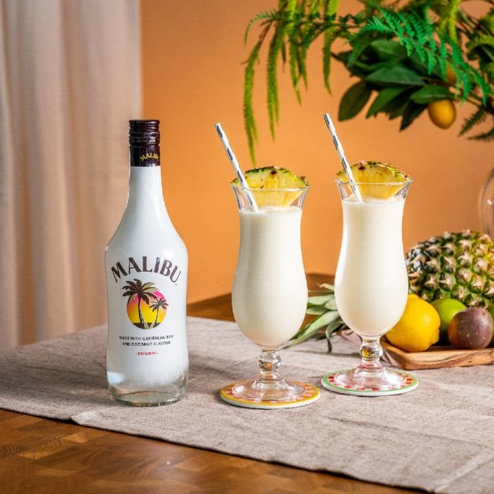 To Slushy Pinã Colada på et bord med en flaske Malibu og frugter