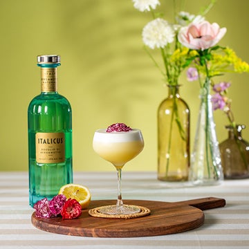 En Italicus Sour cocktail på et bord med en flaske Italicus likør og en buket blomster.
