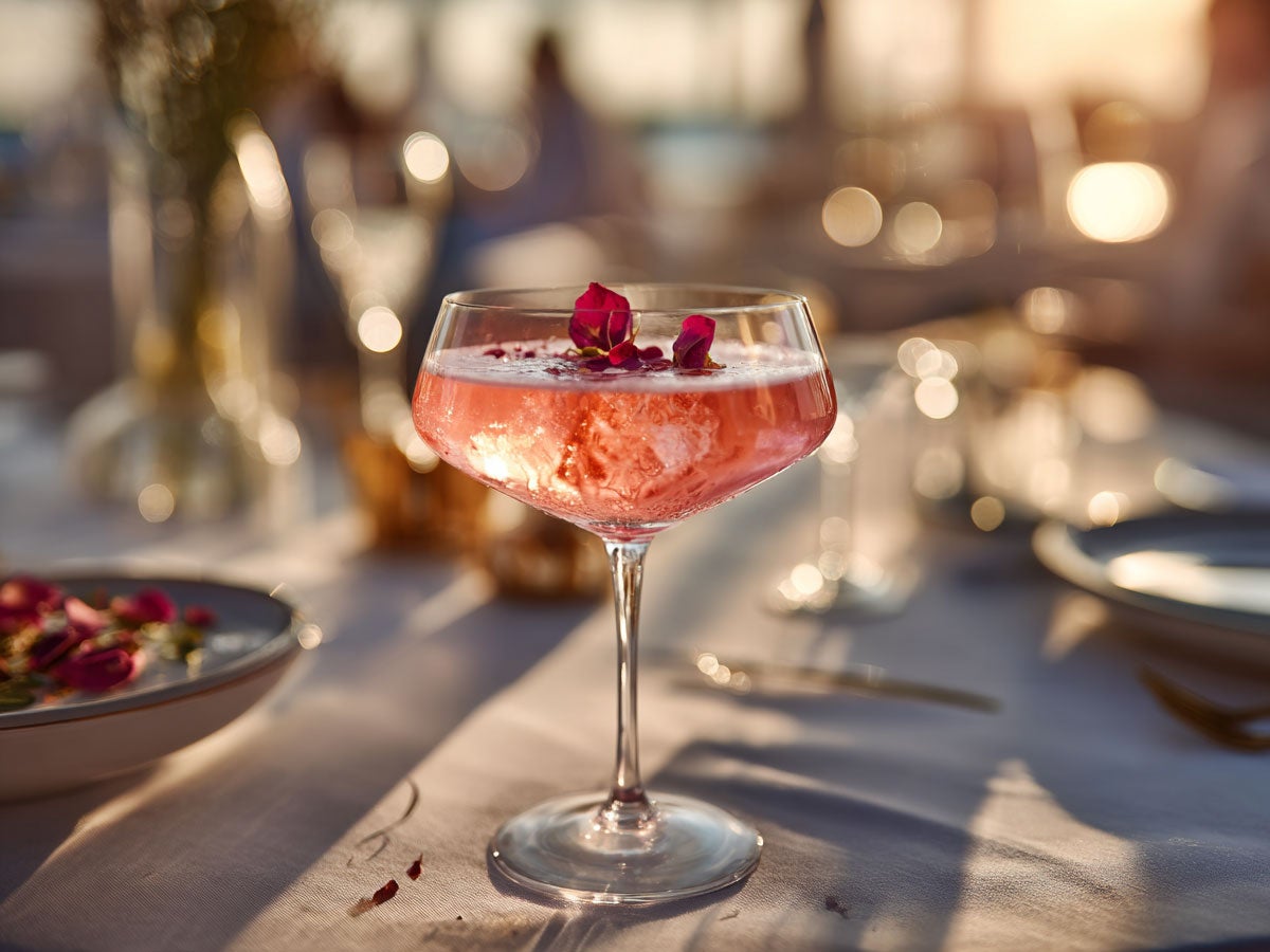 A bright pink drink placed on a set table, garnished with rose petals. Created by AI. Edited by Pernod Ricard Sweden